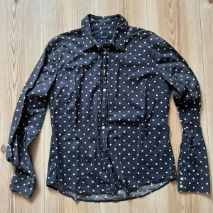 Svart prickig skjorta från Gant - Svart skjorta från Gant med vita prickar, klassisk krage och knäppning framtill. Skjortan har lång ärm och är tillverkad i mjuk bomull. Perfekt för dig som gillar en lekfull men stilren look.