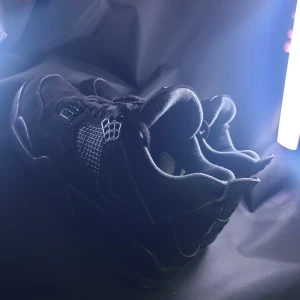 Nike Air Jordan 4 Retro Black Cat - Säljer ett par Nike Air Jordan 4 Retro Black Cat sneakers i helsvart mocka och mesh. Skorna har klassisk Jumpman-logga på plösen och hälen, samt coola detaljer i plast och snörning. Perfekta för dig som gillar stilrena och ikoniska sneakers med streetkänsla.