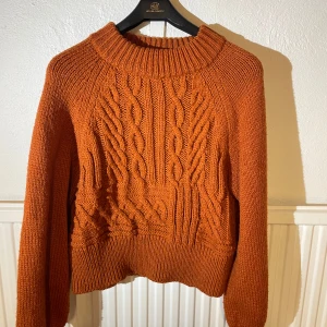 Rostbrun stickad tröja från Monki - Säljer en chunky rostbrun stickad tröja från Monki i storlek M. Tröjan har grovt kabelstickat mönster framtill, bred ribbad krage och muddar vid ärmslut och nederkant. Perfekt för kyliga dagar och supermysig att bära.