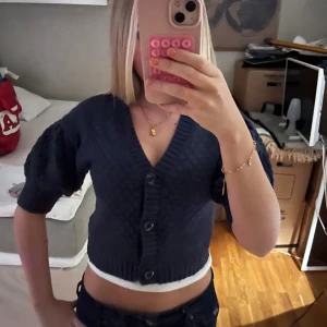 Mörkblå stickad croppad bolerotopp - Säljer en mörkblå stickad bolerotopp med korta puffärmar och djup v-ringning. Toppen har stora knappar framtill och ribbade kanter. Den är croppad och har en snygg strukturstickning som ger en cool look.
