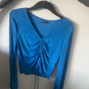 Blå långärmad topp från Gina Tricot - Säljer en blå långärmad topp från Gina Tricot med v-ringning och snygg snörning framtill som ger en rynkad effekt. Toppen är croppad och har en tight passform, perfekt att styla med höga jeans eller kjol.