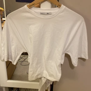 Vit knyt-topp från 157, storlek S - Vit kortärmad topp från 157 i mjuk bomull. Modellen har en enkel rund halsringning och en snygg knytning bak i midjan som ger en trendig och avslappnad look. Perfekt till jeans eller kjol för en clean och fräsch stil.