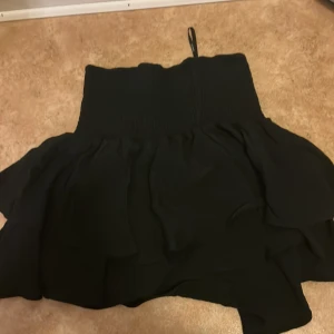 Svart volangkjol från SHEIN - Svart kortkjol från SHEIN med bred smock i midjan och dubbla volanger som ger en snygg och flowy look. Kjolen är enkel att matcha och har en feminin siluett. Perfekt för dig som gillar trendiga och bekväma plagg.