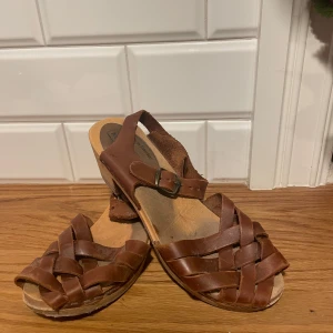 Bruna sandaler från Moheda, stl 39 - Snygga bruna sandaler från Moheda i klassisk svensk stil. Skorna har flätade remmar i skinn, öppen tå och justerbart spänne vid ankeln. Sulan är i trä-look och ger en naturlig vibe. Perfekta för varma dagar och enkel att matcha med sommarens outfits.