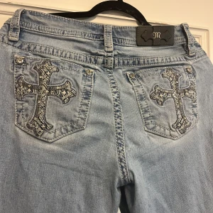 Miss Me ljusblå bootcut jeans stl 30 - Ljusblå jeans från Miss Me med snygga broderade kors på bakfickorna och glittriga detaljer vid fickorna. Modellen har bootcut-ben och klassisk femficksdesign. Perfekta för dig som gillar statement-jeans med unik touch. Jeansen är i bra skick, men har tidigare gått sönder vid sömnen av fickorna men har sytts ihop. Inget som syns från utsidan. Midjemåttet är 42cm rakt över, innerbenslängden 79cm, hela jeans längden är 106cm