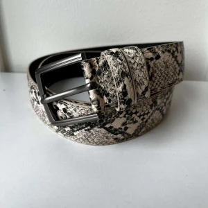 Ormmönstrat bälte - Snyggt bälte med ormmönster i svart, beige och grå toner. Bältet har en klassisk metallspänne och en struktur som påminner om ormskinn. Perfekt accessoar för att lyfta vilken outfit som helst med lite edge. Med extra hål