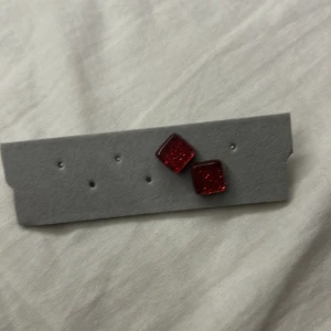 Röda glittriga fyrkantiga örhängen - Snygga örhängen i form av små fyrkanter med röd glittrig yta. Perfekta för dig som vill ha något som sticker ut och ger lite extra färg till din stil. Stift i metall och cool design som passar till många looks.