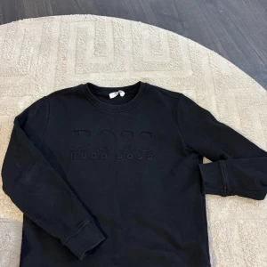 Svart sweatshirt från Hugo Boss - Svart sweatshirt från Hugo Boss med broderad logga framtill. Klassisk rund halsringning, ribbade muddar och mjukt material i bomull. Perfekt för en clean och stilren look. Använd 1-2 gånger därav nyskick!