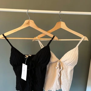 Svart och cream vit  linnetopp från New Yorker - Två snygga linnetoppar från New Yorker, en svart och en cream vit. Båda har smala axelband, knyt framtill och en vågig struktur i tyget. Perfekta för varma dagar och lätta att matcha med jeans eller kjol.