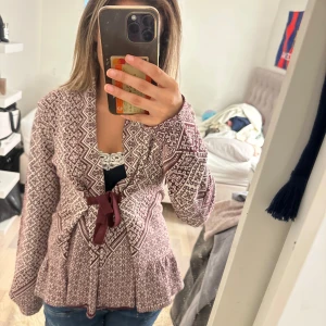 Mönstrad kofta med knyt i vinrött - Säljer en vinröd och vit kofta med snyggt geometriskt mönster och knytband i midjan. Koftan har lång ärm och är stickad i mjukt material, perfekt för kyligare dagar. Passar dig som gillar en mysig och stilren look.