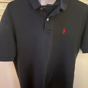 Svart pikétröja Polo Ralph Lauren M - Klassisk svart pikétröja från Polo Ralph Lauren i bomull. Tröjan har korta ärmar, krage och knäppning framtill. Den ikoniska röda broderade loggan syns på bröstet. Perfekt för en clean och stilren look.