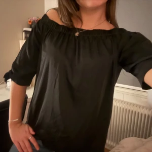 Svart offshoulder blus med rosetter - Svart blus i mjukt, lätt material med offshoulder-modell och trekvartsärmar. Ärmarna har fina rosetter vid avslutet som ger en extra touch. Perfekt att styla med jeans eller kjol för en trendig look.