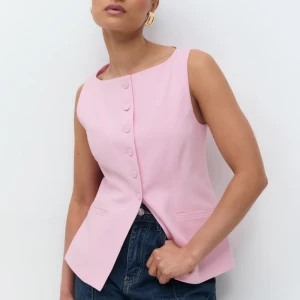 Ljusrosa väst med knappar - Snygg ljusrosa väst med rund hals och raka axlar. Västen har en asymmetrisk knäppning med fem matchande knappar framtill och två diskreta fickor. Materialet är slätt och känns som polyester, perfekt för att ge en fräsch look till jeans eller kjol.