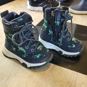 Mörkblå boots med dinosaurier från Gulliver - Säljer ett par mörkblå boots från Gulliver med grönt dinosauriemönster. Skorna har snörning, dragkedja på sidan och grov sula i vitt och svart. Insidan är fodrad för extra värme och komfort.Modell: Lungan Gesunda GTX. Perfekta för kalla dagar och lek utomhus. Finns lite texken på användning efter lek i sandlåda men mest använda da i vagn. Snörena är avklippta och brända för att inte trådas upp. Snörena va alldeles för långa men absolut inget som stör.