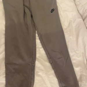 Grå/beiga mjukis sett från Nike - Beige/grå mjukis sett från Nike med elastisk midja och snörning. Byxorna har en enkel design med Nike-logga på låret och är tillverkade i mjukt bomullsmaterial. Hoddien har även en enkel design med Nike loggan på bröstet. Perfekta för chill eller träning.