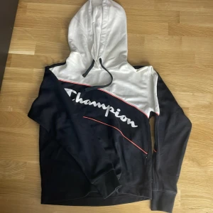 Champion hoodie i svart och vitt - Snygg hoodie från Champion i svart och vitt med röda detaljer och stor logga över bröstet. Tröjan har huva med snörning och långa ärmar. Perfekt för en sportig och avslappnad stil.