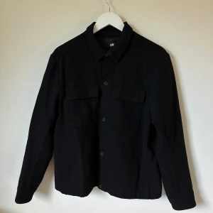 Svart kort rock från H&M i ullmix - Stilren svart kort rock från H&M i en skön ullmix. Jackan har klassisk krage, knäppning framtill och två stora bröstfickor med lock. Perfekt för lager-på-lager och passar till det mesta. Materialet är 54% polyester, 30% ull, 14% akryl och 2% polyamid. 2L6