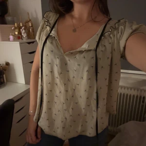 Beige blus med svart knytband - Supersöt beige blus med små svarta mönster och volangdetaljer vid axlarna. Blusen har korta ärmar och ett svart knytband vid halsen som ger en extra touch. Luftig och flowy passform, perfekt till jeans eller kjol.