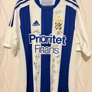 IFK Göteborg signerad matchtröja Adidas 2012-2013 - Blåvit randig fotbollströja från IFK Göteborg med klubbmärke, Adidas-logga och sponsortryck på bröstet. Tröjan har korta ärmar, klassiska Adidas-ränder på axlarna och är signerad av flera spelare. Tillverkad i lätt och ventilerande material.
