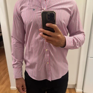 Randig rosa skjorta från Morris - Säljer en snygg långärmad skjorta från Morris med rosa, vita och blå ränder. Skjortan har klassisk krage, knäppning framtill och Morris-logga broderad på bröstet. Perfekt för dig som gillar färg och stilren design.