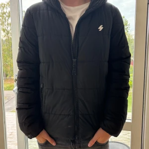 Svart pufferjacka med huva - Svart pufferjacka med huva och dragkedja framtill. Jackan har ett stilrent, quiltat mönster och en liten vit logga på bröstet. Passformen är normal och jackan har långa ärmar. Perfekt för kalla dagar när du vill hålla värmen och se chill ut.