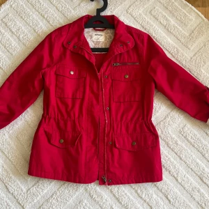 Röd parkajacka från DeFacto - Parkajacka i stark röd färg från DeFacto med fyra fickor framtill, dragkedja och tryckknappar. Jackan har snörning i midjan och långärmade ärmar med mönstrat beige foder. Perfekt för dig som vill ha en cool och praktisk jacka med snygga detaljer.