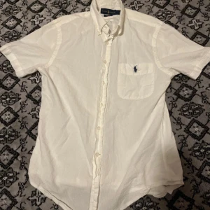 Vit kortärmad skjorta Ralph Lauren - Vit kortärmad skjorta från Ralph Lauren i slim fit med klassisk krage och knappar hela vägen. Tillverkad i 100% bomull med seersucker-struktur och broderad logga på bröstfickan. Perfekt för en clean och fräsch stil.