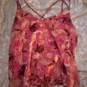 Blommig rosa linnetopp med glitter - Säljer en superfin linnetopp med blommigt mönster i olika rosa och röda nyanser. Toppen har tunna axelband, glittriga detaljer och ett luftigt, flowy tyg som känns lätt på kroppen. Perfekt för dig som vill sticka ut med färg och mönster!
