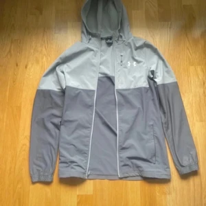 Grå vindjacka från Under Armour - Snygg tvåfärgad grå vindjacka från Under Armour med huva och dragkedja hela vägen. Jackan har lång ärm, diskret logga på bröstet och praktiska fickor. Perfekt för blåsiga dagar och sportiga outfits. Två minimala hål som knappast märks av. Pris diskuteras vid snabb affär.