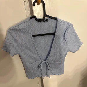 Ljusblå croppad topp från BikBok - Supersöt ljusblå croppad topp från BikBok med korta ärmar, v-ringning och knyt framtill. Toppen har en ribbad struktur och vågiga kanter vid ärmslut och nederkant. Perfekt för dig som gillar en enkel men trendig stil.