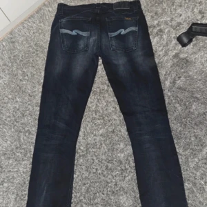  Nudie Jeans 29/32 - Snygga mörkblå jeans från Nudie Jeans med klassisk femficksdesign och ljusblå broderade detaljer på bakfickorna. Jeansen har en rak passform och är tillverkade i slitstarkt denim. Perfekta för dig som gillar stilrena och tidlösa jeans. Tube Lee, sitter som 29 inte 30 i midjan.
