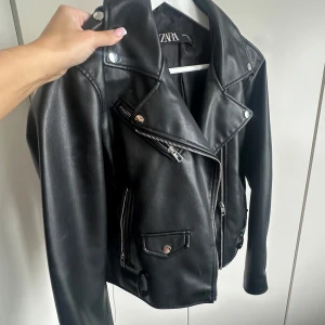 Svart skinnjacka från Zara - Snygg svart skinnjacka från Zara med klassisk bikerstil. Jackan har silvriga dragkedjor, tryckknappar och flera fickor framtill. Materialet är slätt och jackan har en cool, tidlös look med lång ärm och markerad krage. Den är använd några fåtals gånger men har inga defekter! 