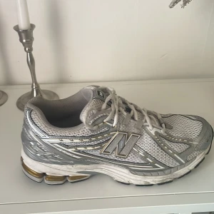 New Balance 2002R silver sneakers - Säljer ett par New Balance 2002R sneakers i silver och vitt med mesh och syntetmaterial. Skorna har coola metallicdetaljer, snörning och en chunky sula med inslag av guld. Perfekta för dig som gillar sportig och trendig stil.