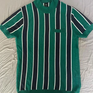 Randig grön t-shirt River Island M - Snygg t-shirt från River Island i storlek M med gröna och marinblå vertikala ränder och vita detaljer. Modellen har korta ärmar, ribbad krage och en liten bröstficka. Materialet känns mjukt och är  bomull. Använd ett par gånger