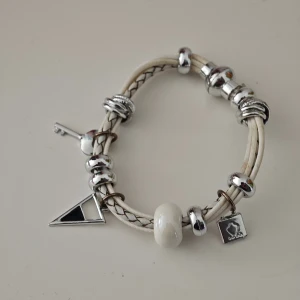 Vitt läderarmband med silverberlocker - Snyggt armband i flätat vitt läder med flera silverfärgade berlocker, bland annat en nyckel, en trekant och en fyrkantig charm. Armbandet har även en stor vit pärlberlock och coola detaljer som ger en unik look.
