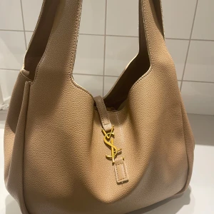 Beige axelväska - Snygg beige axelväska i mjukt, kornigt skinn. Väskan har en stilren design med bred axelrem och ikoniskt spänne i guld framtill. Perfekt storlek för att få plats med allt du behöver och ger en lyxig känsla till din outfit.