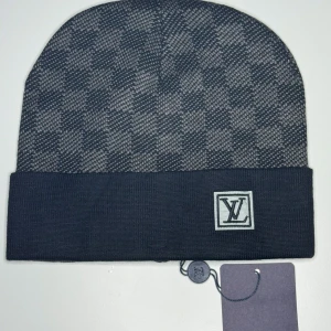 Rutig mössa från Louis Vuitton - Snygg rutig mössa från Louis Vuitton i mörkblått och grått. Mössan har vikt kant med LV-logga framtill och ett klassiskt rutmönster. Perfekt accessoar för att lyfta din stil under kyliga dagar.