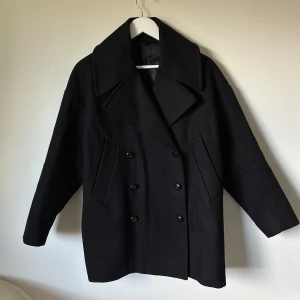 Svart skepparkappa  från Filippa K - Stilren svart skepparkappa från Filippa K, modell Dana Peacoat. Jackan har dubbelknäppning, bred krage och två snedställda fickor framtill. Tillverkad i en ullblandning med polyesterfoder. Perfekt för dig som gillar klassisk och tidlös stil. 3Q13