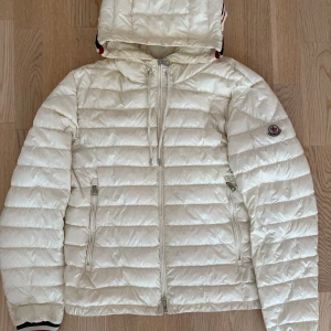 Moncler Giroux - Säljer en snygg vit dunjacka från Moncler med huva och dragkedja. Jackan har quiltad design, Moncler-logga på ärmen och coola röd-blå-vita detaljer på huva och muddar. Perfekt för dig som vill ha en stilren och sportig look under kalla dagar. De är modellen Giroux. Size 2 motsvarar S. För frågor kom pm / Neo 💫