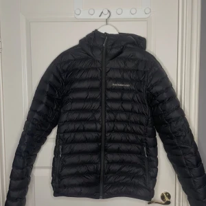 Svart dunjacka Peak Performance M - Svart dunjacka från Peak Performance i storlek M. Jackan har huva, dragkedja framtill och två sidofickor med dragkeda. Jackan är andvänd 1 vinter så inga tecken på andndning. Så gott som ny. Perfekt för kyliga dagar.