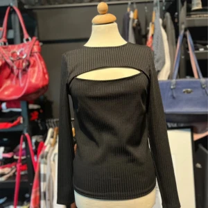 Svart ribbad topp med cut out  - Svart långärmad topp från Gina Tricot med ribbad struktur och cool cut out-detalj över bröstet. Slimmad passform som sitter snyggt och ger en trendig look. Perfekt för dig som vill sticka ut lite extra.