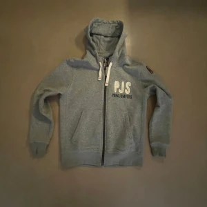 Grå hoodie från Parajumpers  - Snygg grå hoodie från Parajumpers med dragkedja, broderad logga på bröstet och patch på ärmen. Mjuk fleece på insidan, huva med snörning och coolt tryck på huvan. Perfekt för kyliga dagar och chill outfits. 