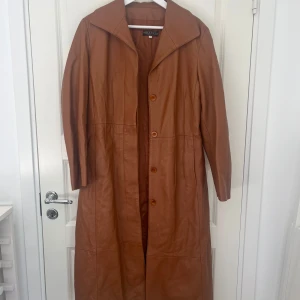 Brun lång skinnkappa från Vera Pelle - Säljer en snygg lång kappa i äkta brunt skinn från italienska Vera Pelle. Kappan har klassisk krage, knäppning med knappar framtill och raka långa ärmar. Perfekt för dig som vill ha en tidlös och cool look med vintagekänsla.