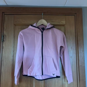 Rosa hoodie med dragkedja - Säljer en ljusrosa hoodie med svart dragkedja och huva. Tröjan har långa ärmar, fickor framtill och en liten logga nere vid fållen. Perfekt för en avslappnad och chill stil.