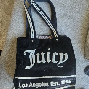 Juicy Couture svart axelväska  - Juicy Couture axelväska i svart sammet med stora vita broderade logotexter på framsidan och handtag. Insidan har ett mönstrat foder med Juicy Couture-tryck, dragkedjefack och en silverfärgad liten necessär med logga. Äkta och i fint skick hittar inga skavanker :) 