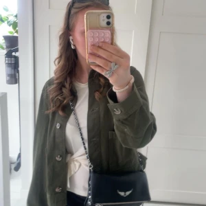 Olivgrön jacka med knappar - Säljer en olivgrön overshirt med klassisk krage och stora fickor framtill. Jackan har en loose passform och stängs med knappar. Materialet är bomull och den har långa ärmar. Perfekt att slänga över en t-shirt eller topp för en chill look.