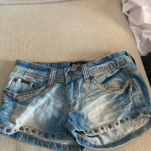 Blå jeansshorts med nitar från ONLY - Snygga blå jeansshorts från ONLY i tvättad denim med slitningar och coola nitar längs nederkanten. Klassisk femficksmodell med orange kontrastsömmar och knappstängning. Perfekta för en chill sommarlook.