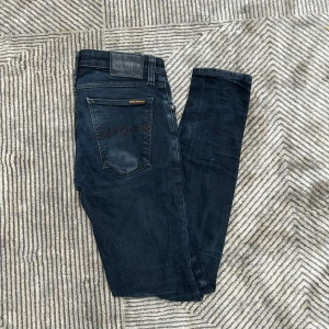 Mörkblå jeans från Nudie Jeans - Säljer ett par mörkblå skinny jeans från Nudie Jeans i storlek 29/32. Jeansen har klassisk femficksdesign, svarta sömmar och en diskret tvättad look. Materialet är stretchigt och sitter tight hela vägen ner. Perfekta för dig som gillar en smal siluett. Fixar hål i skrevet 