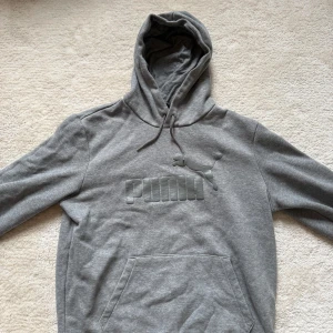 Grå hoodie från Puma med logga - Säljer en grå hoodie från Puma i mjuk bomullsblandning. Tröjan har stor huva med snörning, klassisk känguruficka och Puma-logga i relief framtill. Perfekt för chill dagar eller när du vill ha en enkel streetstyle-look.