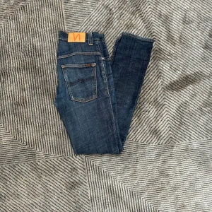 Mörkblå jeans från Nudie - Säljer ett par mörkblå skinny jeans från Nudie med klassisk femficksdesign och orange logga på bakfickan. Jeansen har smal passform, snygga slitningar och är tillverkade i stretchig bomullsdenim för extra komfort.
